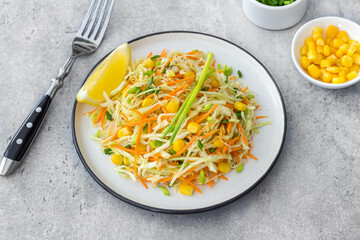 Fresh healthy cabbage salad with carrot, corn and scallions garnished with lemon on white plate on a gray stone background