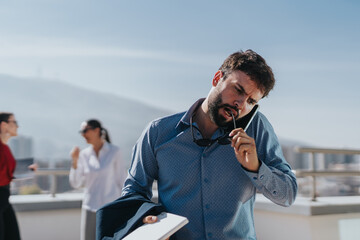 A focused businessman multitasks outdoors, holding a phone and chewing on his glasses. Colleagues engage in the background, depicting a busy professional environment.