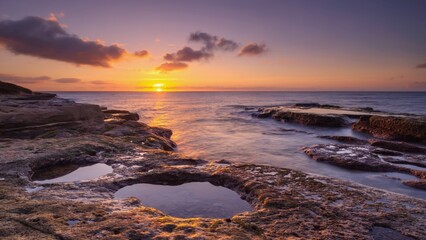 A serene beach scene at sunset with a clear sky and calm ocean. The rocks create interesting patterns on the shore.