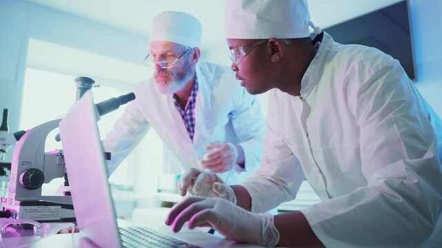Two professional scientists wearing lab coats, bouffant caps, and safety glasses, collaborate on research, using a microscope and laptop in a brightly lit laboratory