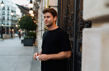 Stylish young man in black tee stands calmly with wireless earbuds in, holding charging case,...