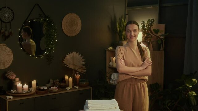 Portrait of young masseuse smiling and folding her arms while posing for camera in authentic spa room