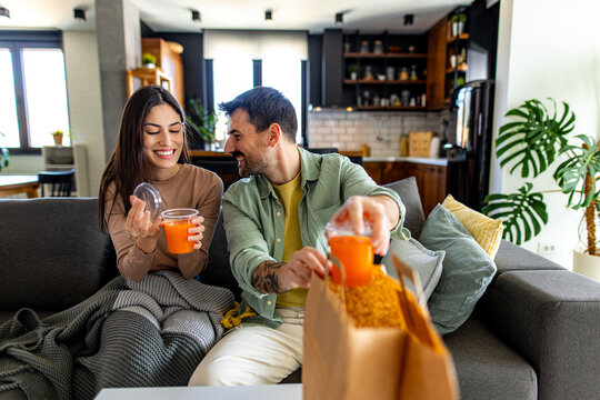 Young couple smiling and unpacking takeaway food and drinks while relaxing on the sofa in their cozy living room, enjoying each other’s company