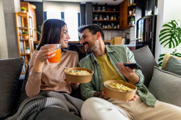 Happy couple eating pasta from takeaway boxes and watching television using a remote control,...