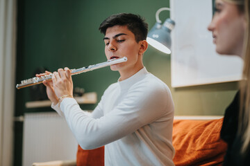 A young man dressed in a white shirt practices playing the flute indoors, immersing in music while...