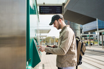 Young man buying transport pass at modern tram train station using mobile phone app and ticket...