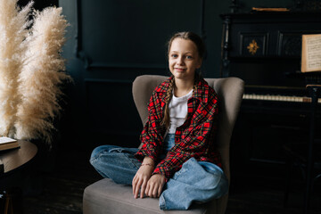Adorable preteen girl sitting cross-legged on armchair, braided hair framing happy expression....