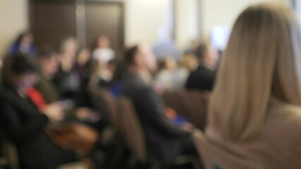 Woman is sitting in a room with a group of people. The room is filled with chairs and people are sitting in them. The woman is looking at the camera