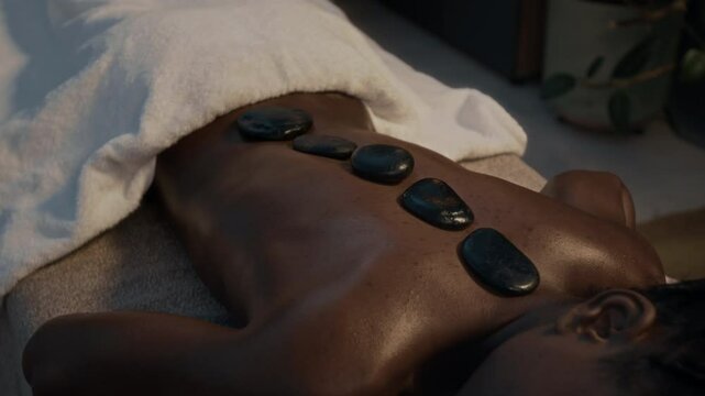 Close-up shot of unrecognizable African American woman lying face-down on massage table, draped in white towel, receiving hot stone massage in serene spa