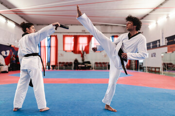 A male martial artist in a white uniform and black belt performs a high kick toward a target pad held by a female partner inside a red-curtained training hall