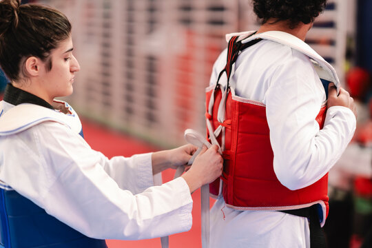A female martial artist wearing a blue chest protector helps tie the red chest guard of her male partner in a training gym with red and blue mats and background mural