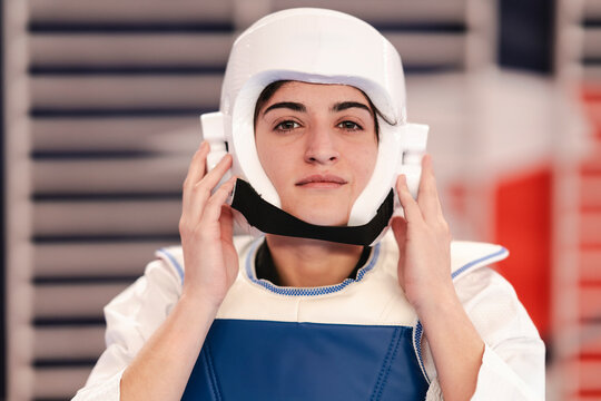 A focused female martial artist looks directly ahead while wearing a white helmet and blue chest protector during taekwondo preparation inside a gym with padded walls