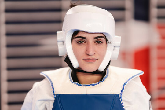 A focused female martial artist looks directly ahead while wearing a white helmet and blue chest protector during taekwondo preparation inside a gym with padded walls