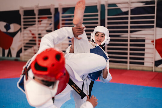 A female martial artist in blue protective gear prepares to block a high kick from her male opponent during a sparring session on a red and blue mat inside a gym