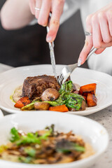 A close-up of a person's hands cutting into a tender piece of meat on a plate, accompanied by colorful vegetables. A second dish is slightly visible in the background.

