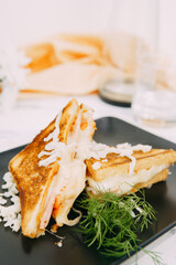 Close-up of toasted sandwiches with cheese filling and garnish, arranged on a dark plate. A yellow napkin, a transparent jug, and a glass decorate the light background.