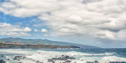 Obraz premium Rocky coast with waves, town, and cloudy sky, Sao Miguel, Azores, Portugal