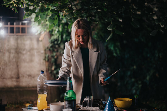 A woman engages in preparation activities outdoors under the evening lights, with a table set up with bottles, dishes, and tools, showcasing a serene and focused moment in a natural setting.