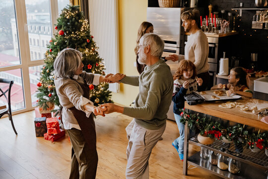 White grandparents dance while their White school-aged grandson and granddaughter eat baked cookies from a tray on the island while their parents dance next to them, in a kitchen with a Christmas tree - Powered by Adobe