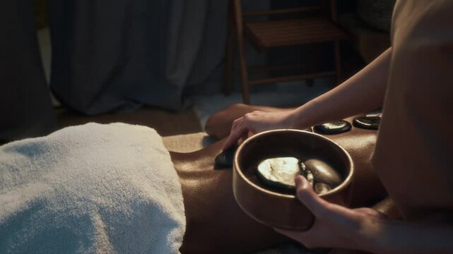 Unrecognizable masseuses hands placing hot stones from bowl onto back of African American woman, recieving massage in spa