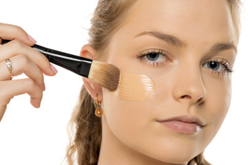 Close-up of young woman applying liquid foundation with makeup brush on cheek, showing smooth application process against white background