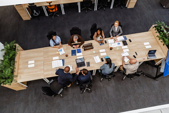 An overhead view of a long table in an office room, where a diverse group of colleagues sit and have a meeting, all dressed in suits and discussing papers