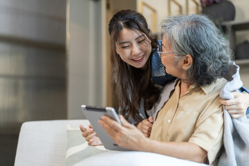 Asian senior mother and daughter smiling and talking using tablet browsing social media, shopping online, watching movies or series, enjoying entertainment digital connection and bonding at home