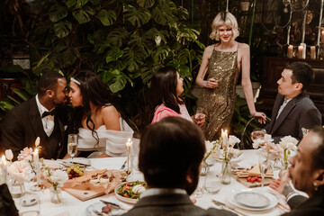 A group of diverse guests dressed elegantly gathers at a dinner party in an indoor setting filled...