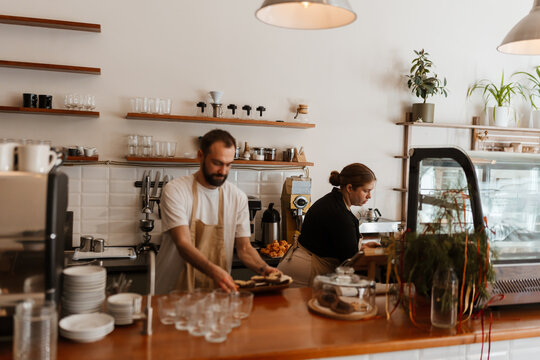 An overweight Caucasian female barista in her mid-20s, with an apron over her clothes, cleaning behind the counter of a cozy coffee shop as her mid-30s White male colleague works beside her