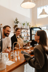 A view from the side of a White male barista in his mid-30s, taking an order from a White businesswoman in her late 20s, as his overweight White female colleague stands beside him, in a cozy caf?