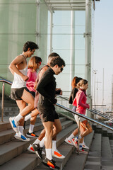 A side view of a multiethnic group of friends in their 20s in athletic clothing and sneakers, running down concrete stairs with a modern glass building and a marina with sailboats in the background. © Drobot Dean
