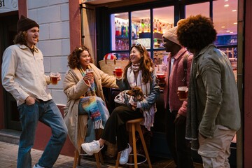 A young Asian woman in her late 20s, with her dachshund dog in her lap, sitting on a stool outside of a bar, on the street, drinking and chatting with her diverse friends, in the evening