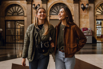 In a train station with tiled flooring, a smiling White lesbian couple in their 20s, a White blonde...