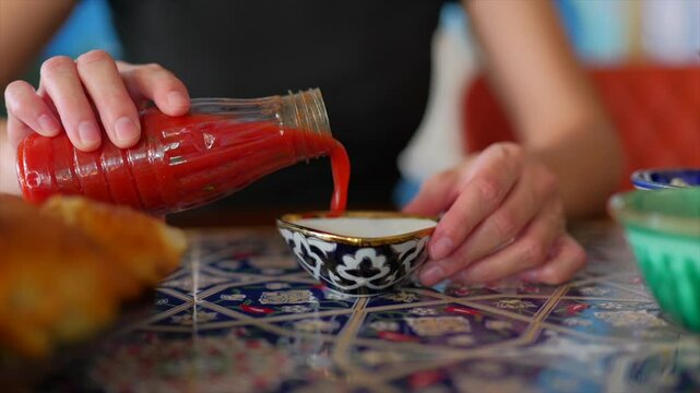 Woman pours red sauce into traditional bowl on vibrant Uzbek table.