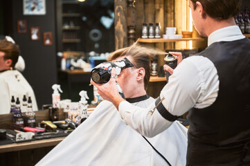 Professional hairdresser using hairdryer and hairspray for styling. Young customer getting new hairstyle. Stylish barber during process.