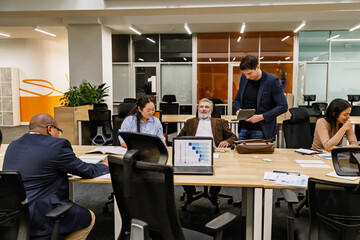 A White elder male office worker is surrounded by a multinational group of colleagues, collaborating in a modern office space with laptops, books, and a data presentation on the table.