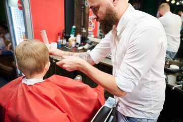 Barber using comb and shaver to cut hair. Professional hairdresser shaving little kid's nape.