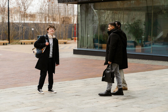 A White young man with curly hair, glasses and headphones, a Black young man, and an Indian young man are casually talking outside during their internship in an urban setting.