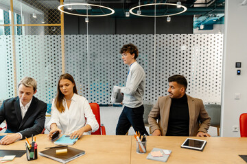 In a business room, three colleagues sit at a table, and behind them, their young White male colleague passes with papers in his hands. Concept of multitasking in the office, preparing for a meeting.
