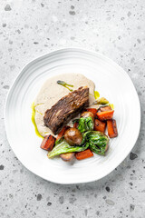 A succulent piece of beef served with roasted vegetables, including carrots and potatoes, drizzled with a creamy sauce on a stylish white plate, creating a hearty meal.
