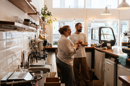 A White man and woman stand behind the counter of a modern cafe, smiling and talking during renovation. Surrounded by coffee equipment, clean dishes, and shelves, they prepare the workspace.