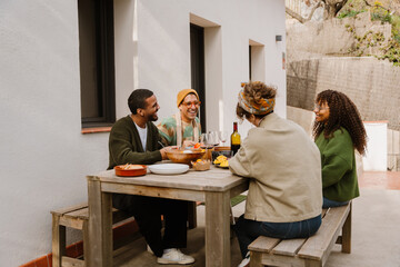 A multiracial group of adult men and women enjoys food and wine while seated at a wooden table outdoors. They smile and engage in conversation in a modern courtyard or terrace during daylight.