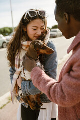 An adult Asian woman holds a dachshund dog while talking to a Black adult man on a city street. Both are dressed warmly in winter clothing and interacting outdoors on a sunny day.