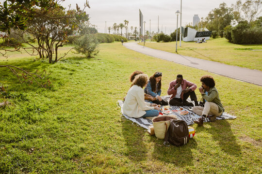 A group of young multiracial friends is having a picnic on a blanket in a city park, sitting on the grass, eating and drinking together on a sunny day with food, backpacks, and snacks around them.