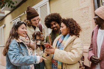 A group of multiracial friends standing on a city street looking at a smartphone together, with one man holding a small dog, all wearing winter jackets and scarves during a casual walk outdoors.