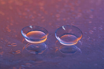 Pair of contact lenses and water drops on purple gradient mirror surface, closeup