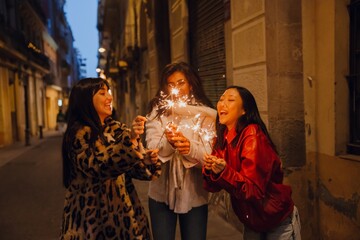 On a city street at dusk, three young women hold lit sparklers in their hands, lighting them against each other and enjoying it. The concept of a street party, mischief, happiness.