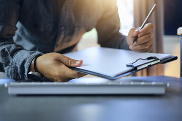 Office worker's hand holding clipboard and writing with pencil on paper to record project progress...