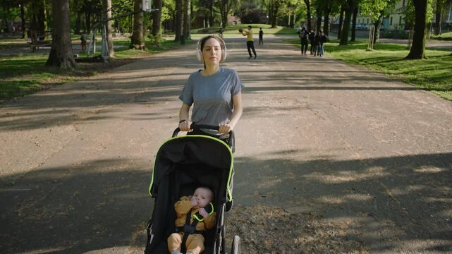 A young sporty woman walks in the park with her son in the running board. Mom is happy to do sports in the park with her newborn baby on a sunny day. Concept of power, fitness, family