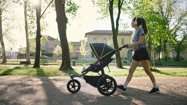 A young sporty woman walks in the park with her son in the running board. Mom is happy to do sports in the park with her newborn baby on a sunny day. Concept of power, fitness, family